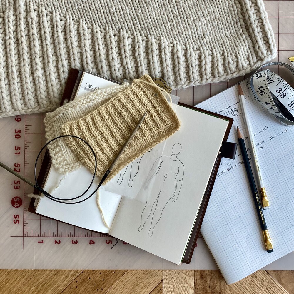  A table shows a number of in-progress making tools: a notebook with a hand-drawn plus-size croquis, some gauge swatches, a pair of knitting needles, a cutting mat, pencils and graph paper, a measuring tape, and the bottom hem of a sweater that is mo… 