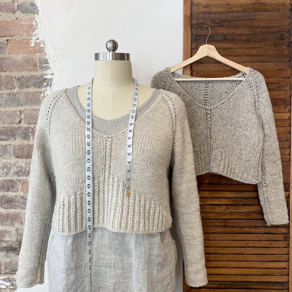  A size 22 dress form displays a 54” cropped sweater (Ursa) over a linen tunic. A measuring tape is draped around the neck of the dress form and a smaller sample is hanging on the wall behind. 